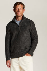 A man wearing a dark charcoal gray textured quarter-zip sweater over a light gray t-shirt and cream-colored pants, standing against a solid light gray background.