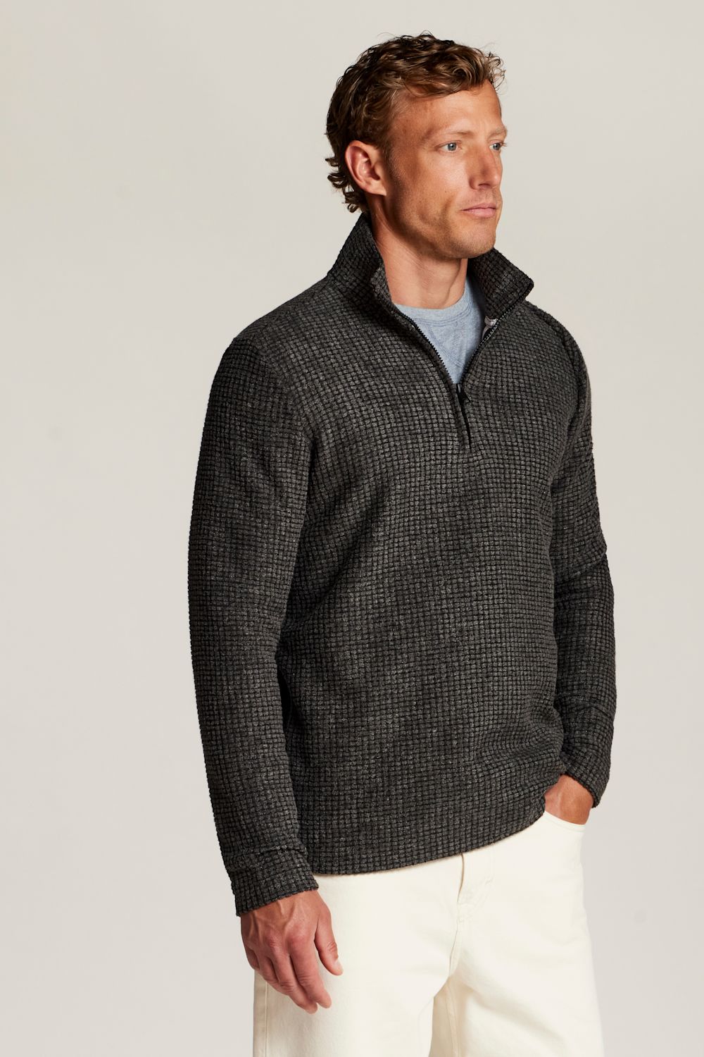 A man wearing a dark charcoal gray textured quarter-zip sweater over a light blue t-shirt and off-white trousers, standing against a plain light gray studio background.