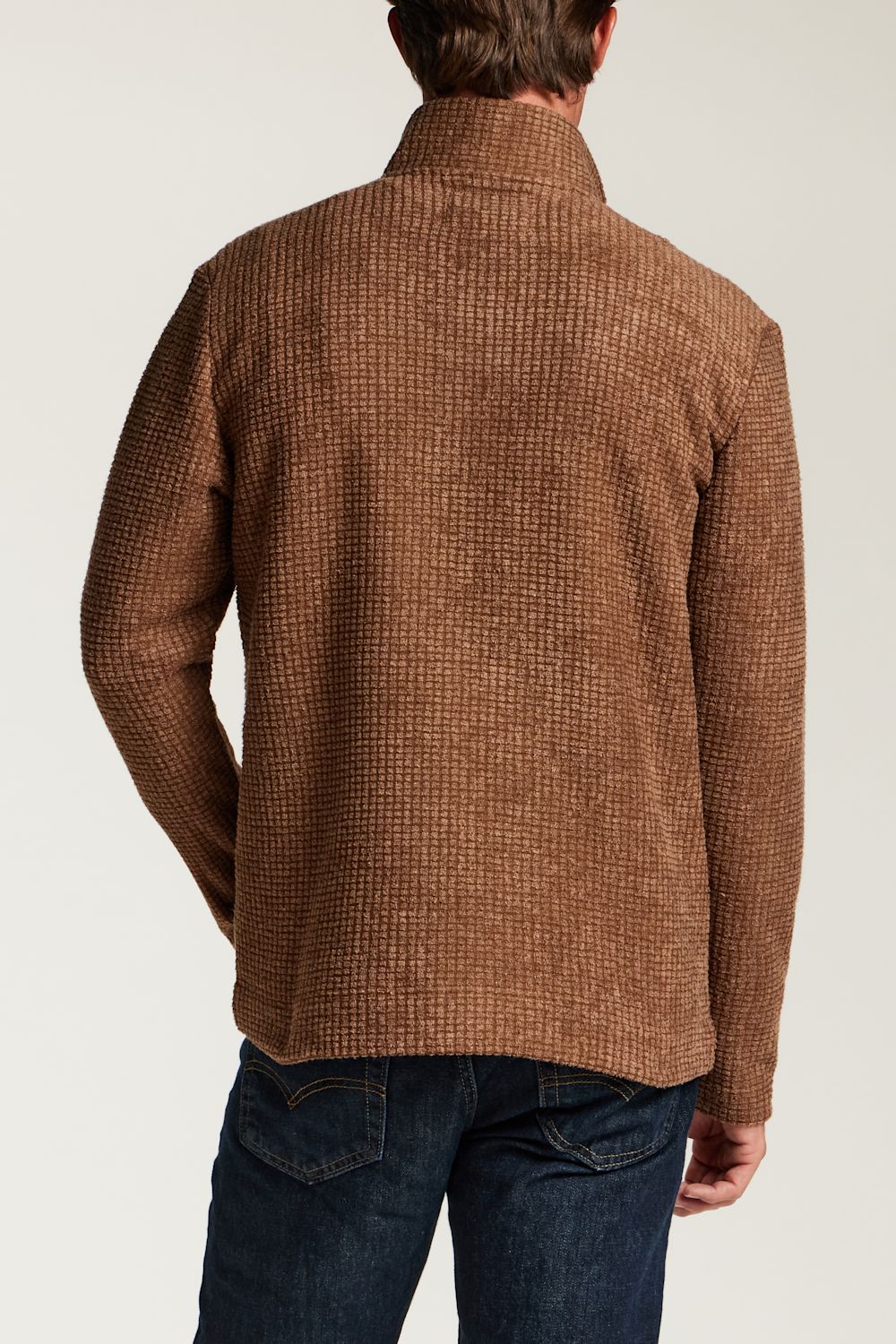 A man viewed from the back wearing a brown textured knit sweater and dark blue jeans against a light gray studio background.