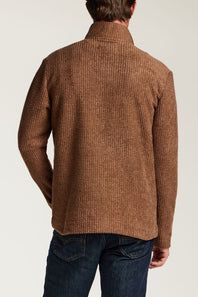 A man viewed from the back wearing a brown textured knit sweater and dark blue jeans against a light gray studio background.
