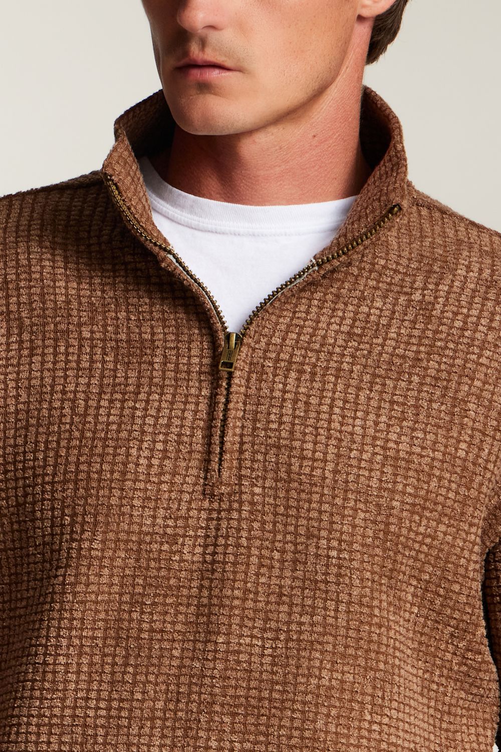 Close-up of a man wearing a brown textured quarter-zip pullover sweater over a white t-shirt, against a light beige background.
