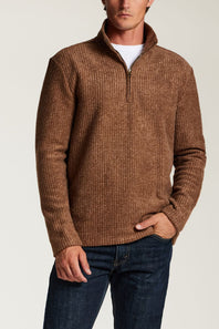 A man wearing a brown textured quarter-zip sweater over a white t-shirt and dark blue jeans, photographed against a light gray studio background.