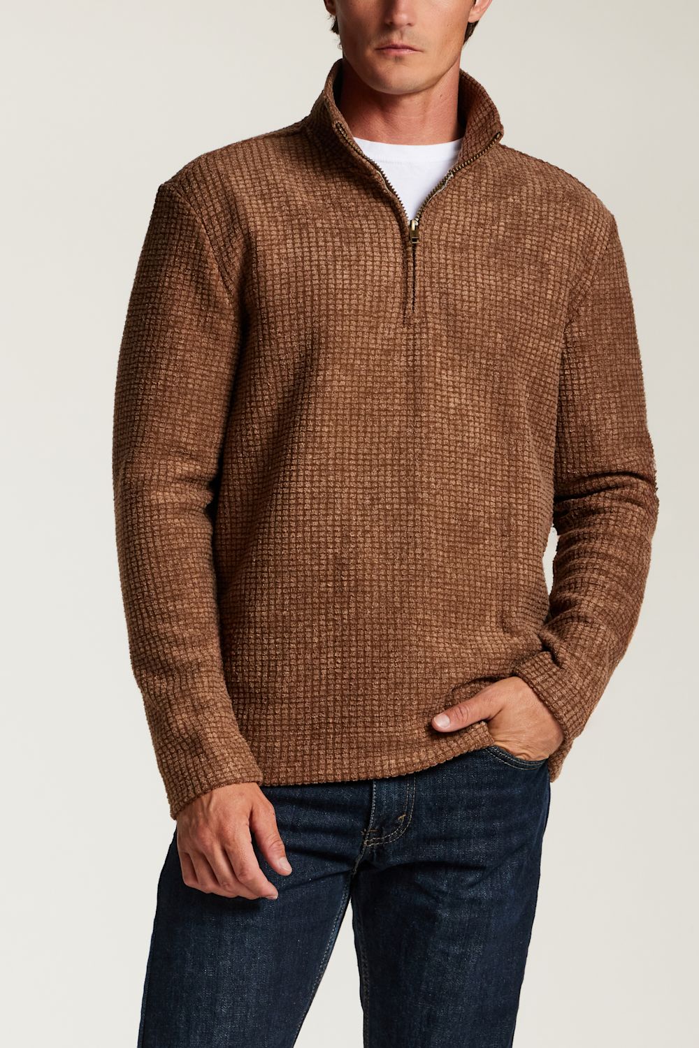 A man wearing a brown textured quarter-zip sweater over a white t-shirt and dark blue jeans, photographed against a light gray studio background.