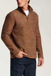 A man wearing a brown textured quarter-zip pullover sweater with a stand collar, paired with dark blue jeans and a white undershirt, shown from the chest down against a light gray studio background.