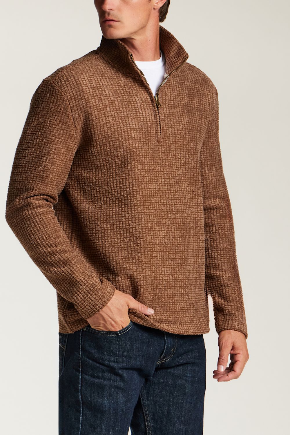 A man wearing a brown textured quarter-zip pullover sweater with a stand collar, paired with dark blue jeans and a white undershirt, shown from the chest down against a light gray studio background.