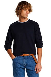 A man with curly light brown hair wearing a dark navy blue long-sleeve crewneck sweater, blue jeans, and a patterned orange-brown belt, photographed against a solid white background.