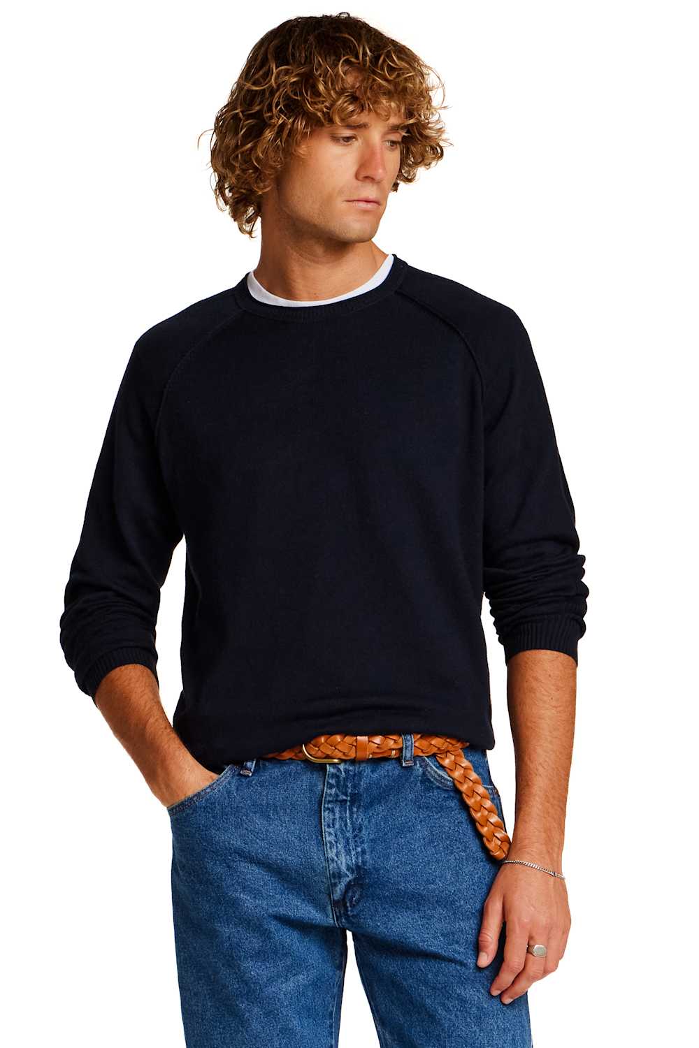 A man with curly light brown hair wearing a dark navy blue long-sleeve crewneck sweater, blue jeans, and a patterned orange-brown belt, photographed against a solid white background.