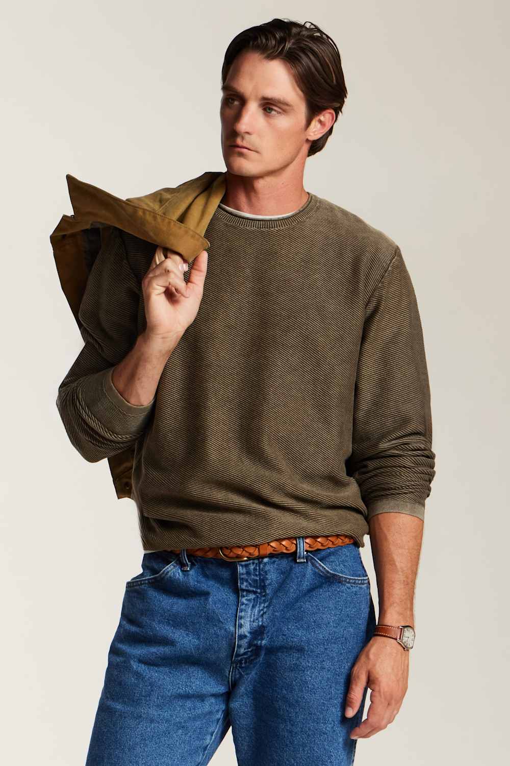 A man wearing an olive green crewneck sweater, blue jeans, and a brown belt, holding a brown jacket over his shoulder against a light gray studio background.