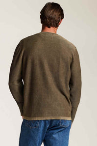 A man viewed from behind wearing an olive green long-sleeve knit sweater and blue jeans against a plain light background.