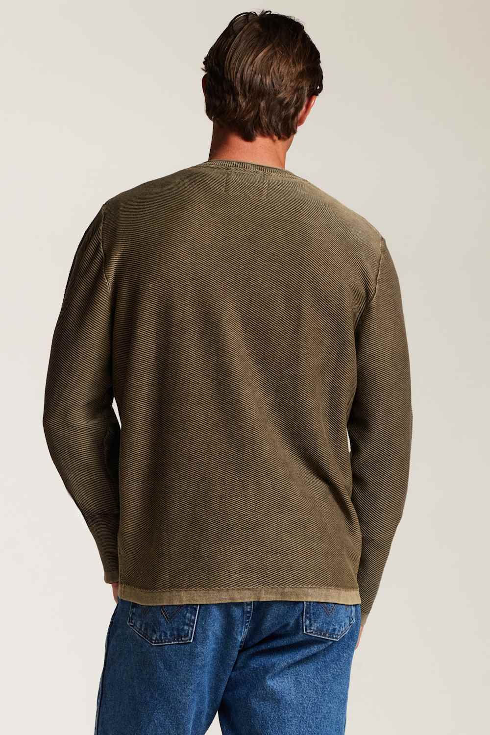 A man viewed from behind wearing an olive green long-sleeve knit sweater and blue jeans against a plain light background.