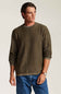 A man wearing a long-sleeve crewneck sweater in olive green, paired with blue jeans, standing against a light beige studio background.