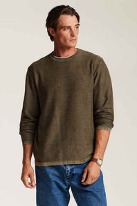 A man wearing a long-sleeve crewneck sweater in olive green, paired with blue jeans, standing against a light beige studio background.
