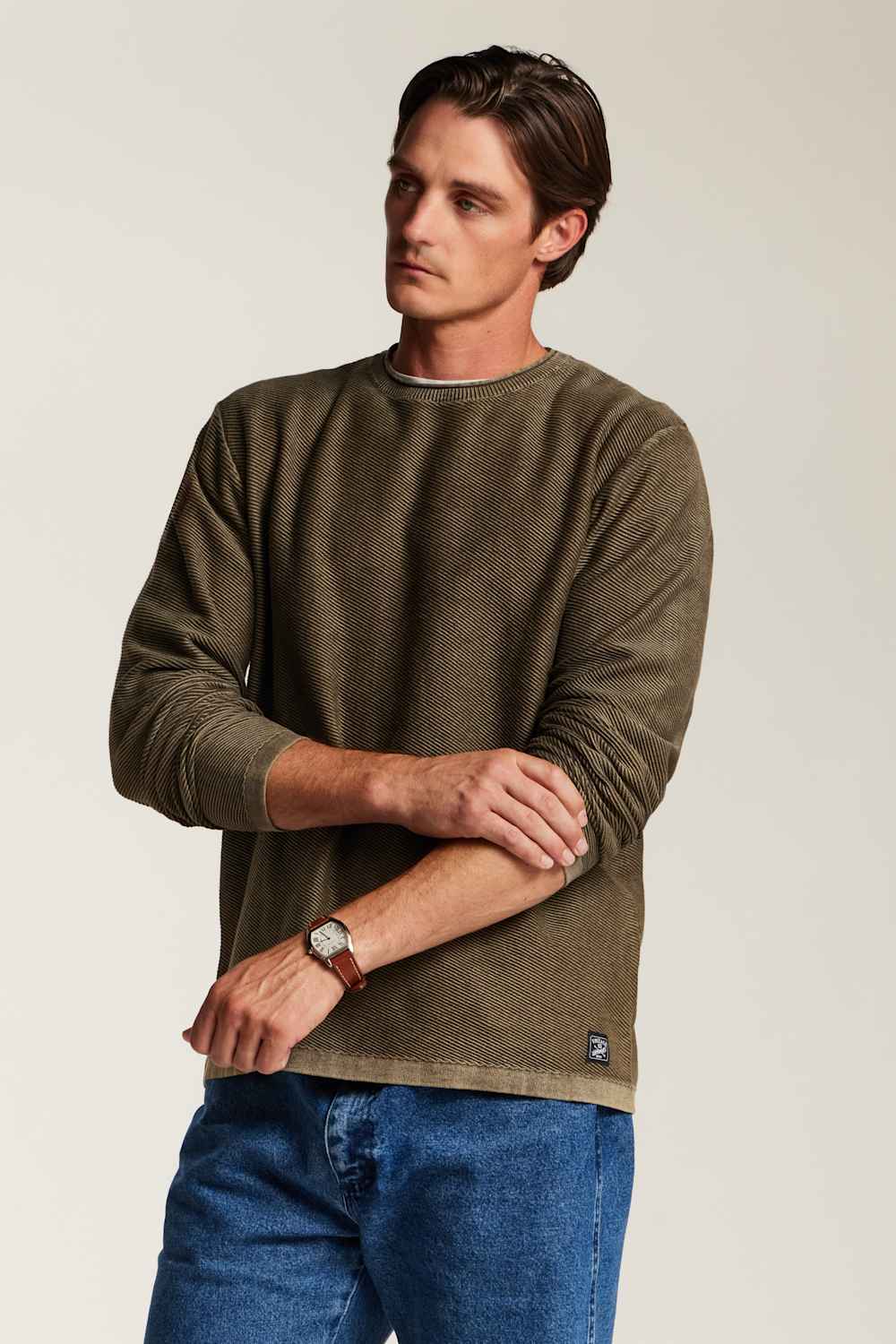 A man wearing a muted olive-brown long-sleeve crewneck sweater and blue jeans, standing against a plain light beige studio background.