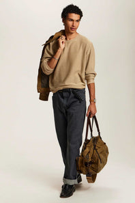 A male model wearing a beige crewneck sweater, dark gray jeans, and black shoes, styled with a brown jacket draped over his shoulder and a brown canvas duffel bag, set against a plain light beige studio background.