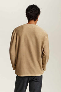 A man viewed from behind wearing a beige long-sleeve crewneck sweater and dark jeans, standing against a plain light gray studio background.