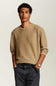 A man wearing a beige crewneck sweatshirt, dark gray jeans, and a black woven belt, standing against a light gray studio background.