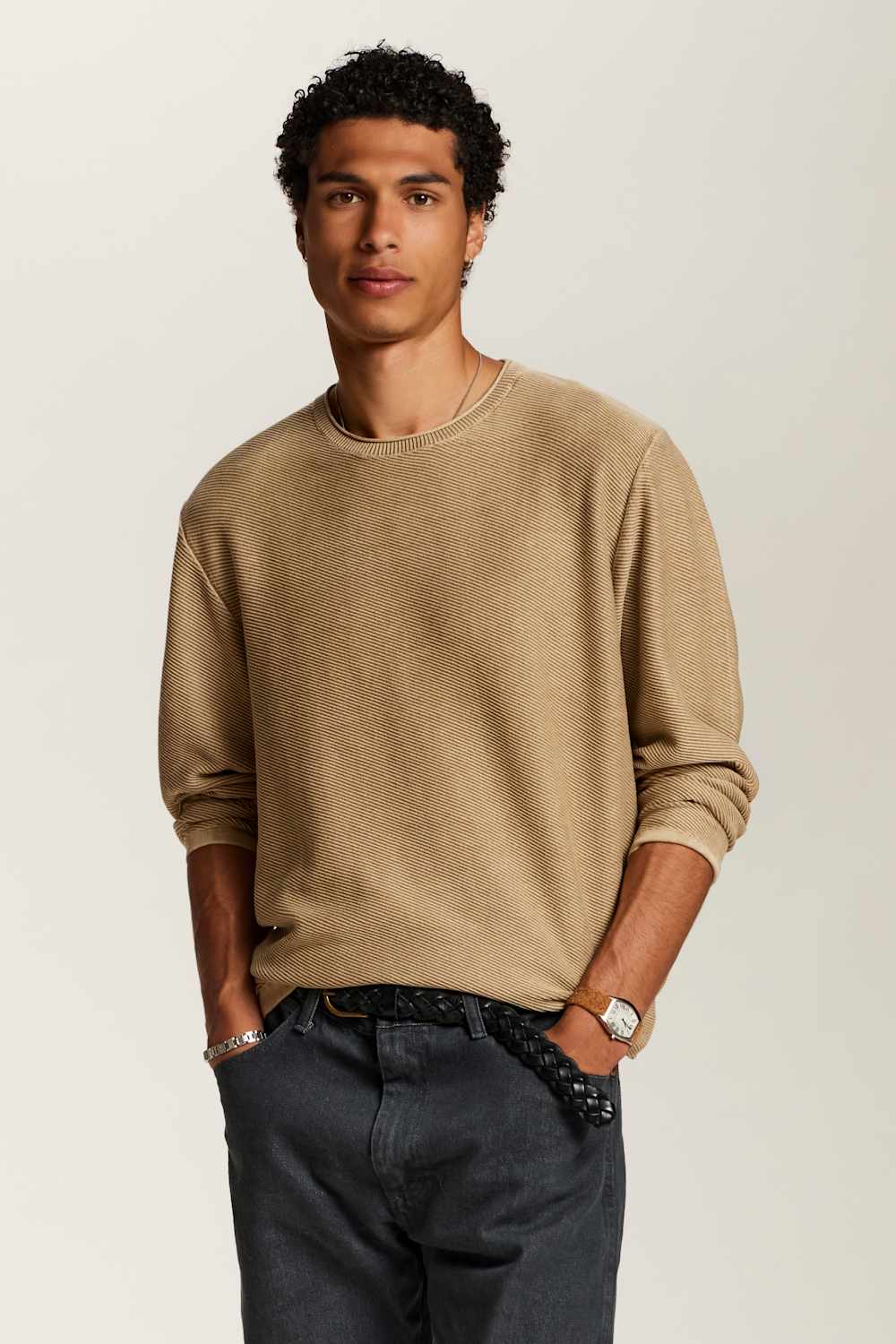 A man wearing a beige crewneck sweatshirt, dark gray jeans, and a black woven belt, standing against a light gray studio background.