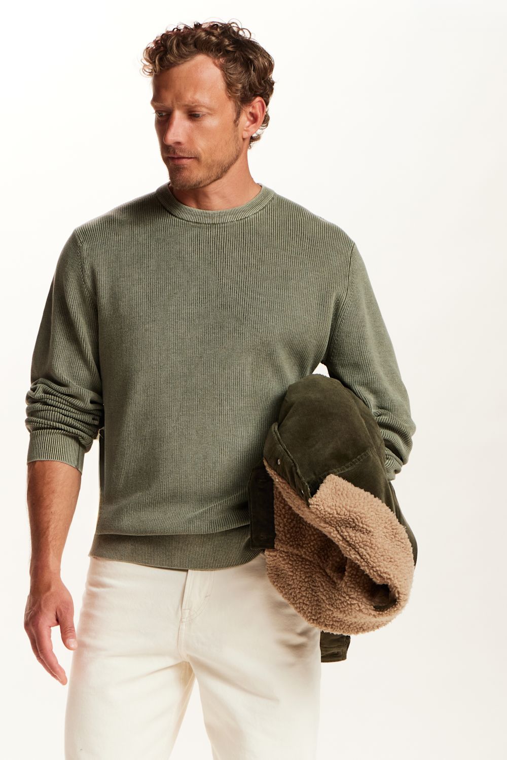 A man wearing an olive green crewneck sweater and off-white trousers, holding a dark green jacket with a beige sherpa lining against a plain white background.