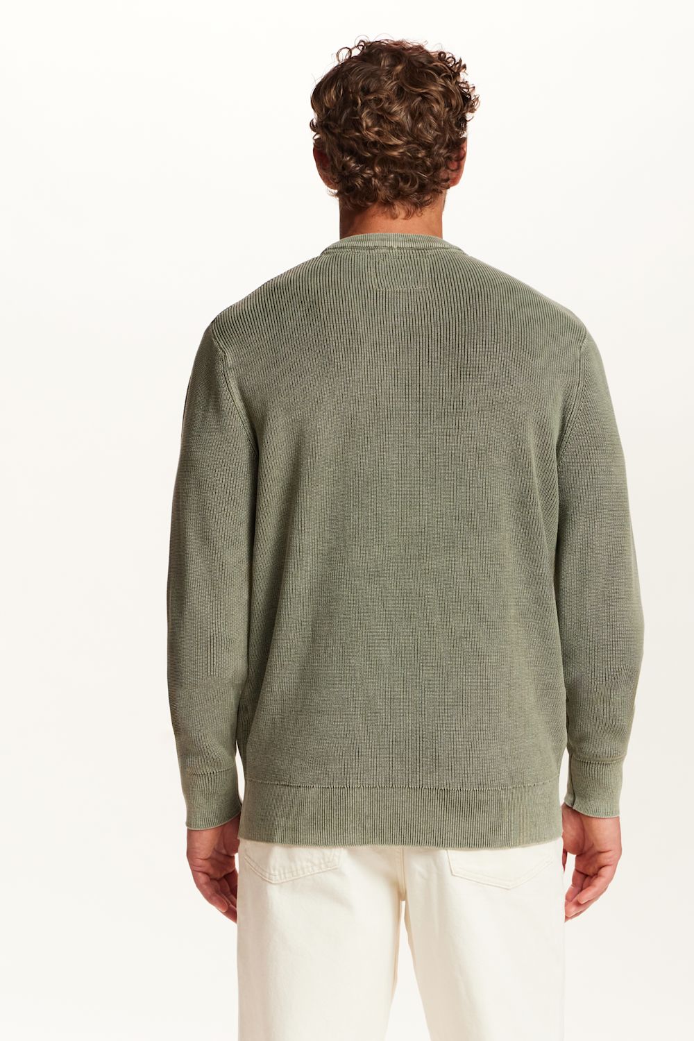 A man viewed from behind wearing a sage green knit sweater and off-white trousers, photographed against a clean white studio background.