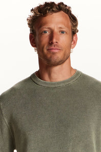 A close-up shot of a man wearing a ribbed knit crewneck sweater in a muted sage green color, set against a plain white background.