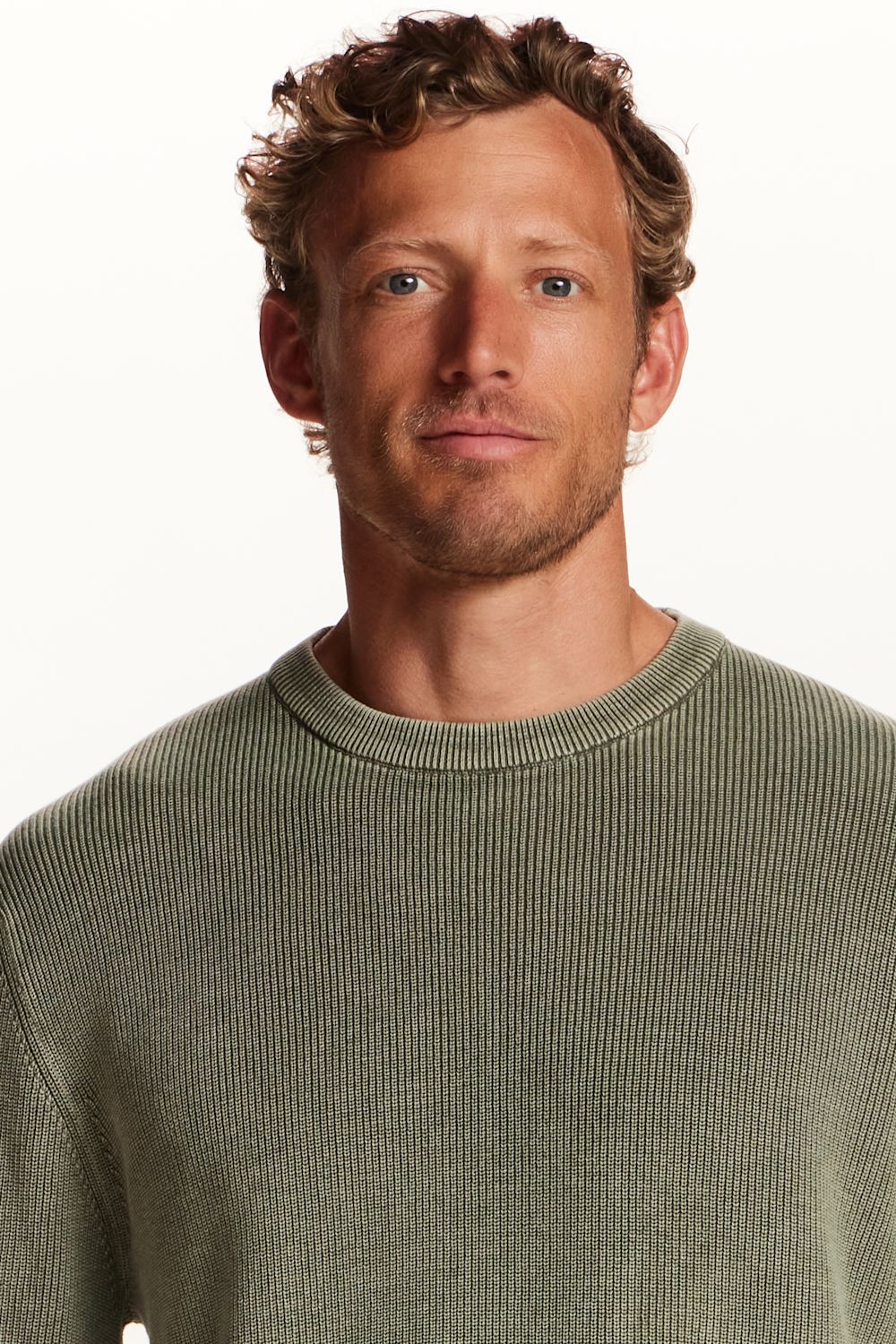 A close-up shot of a man wearing a ribbed knit crewneck sweater in a muted sage green color, set against a plain white background.