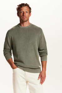 A man wearing a muted sage green crewneck sweater and off-white trousers, standing against a plain white studio background.