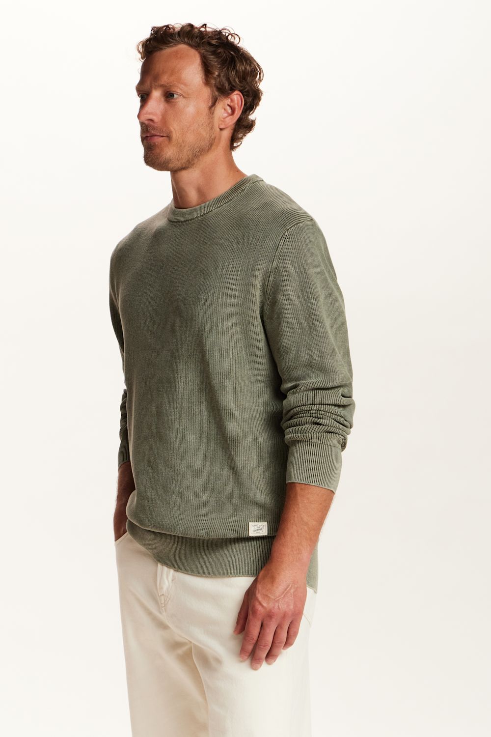 A man wearing a sage green crewneck sweater and off-white trousers, photographed from the side against a clean white studio background.