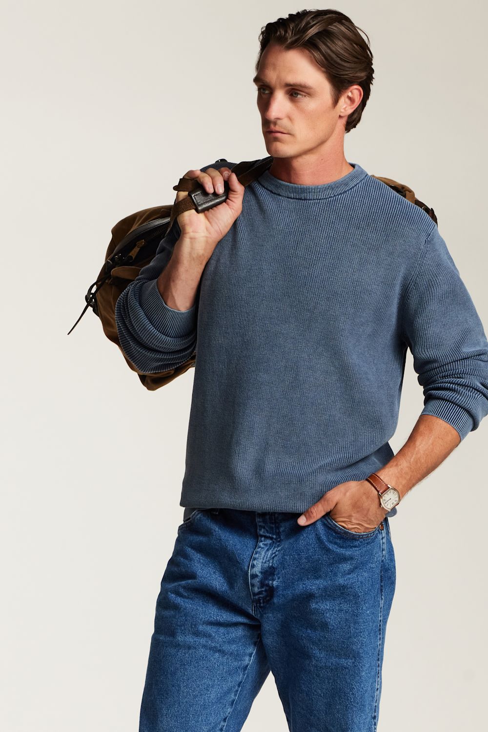 A man wearing a heathered slate blue crewneck sweater and blue jeans, carrying a brown backpack over his shoulder against a light beige studio background.