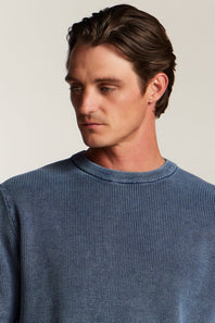 A man wearing a blue ribbed knit crewneck sweater, shown from the chest up against a plain light beige background.
