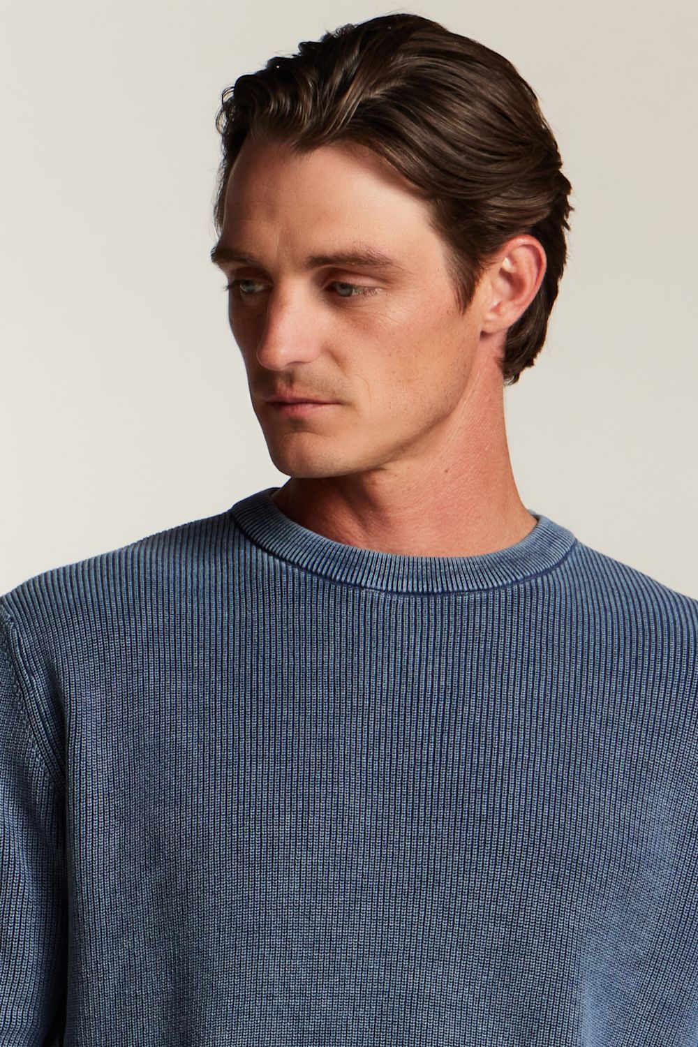 A man wearing a blue ribbed knit crewneck sweater, shown from the chest up against a plain light beige background.