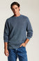 A man wearing a slate blue crewneck sweatshirt and blue jeans, standing against a light beige studio background.