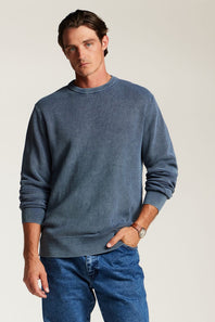 A man wearing a slate blue crewneck sweatshirt and blue jeans, standing against a light beige studio background.
