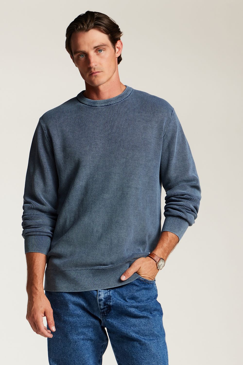 A man wearing a slate blue crewneck sweatshirt and blue jeans, standing against a light beige studio background.