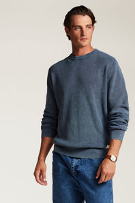 A man wearing a slate blue crewneck sweatshirt and blue jeans, standing against a plain light beige studio background.
