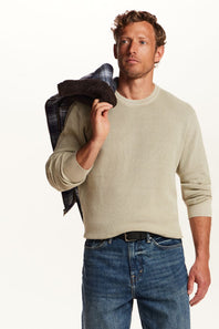 A man wearing a light beige crewneck sweater and blue jeans, holding a dark plaid jacket over his shoulder against a plain white background.