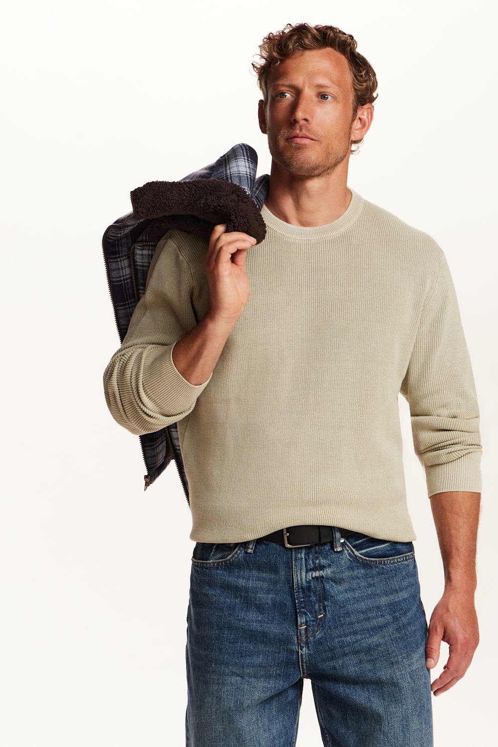 A man wearing a light beige crewneck sweater and blue jeans, holding a dark plaid jacket over his shoulder against a plain white background.