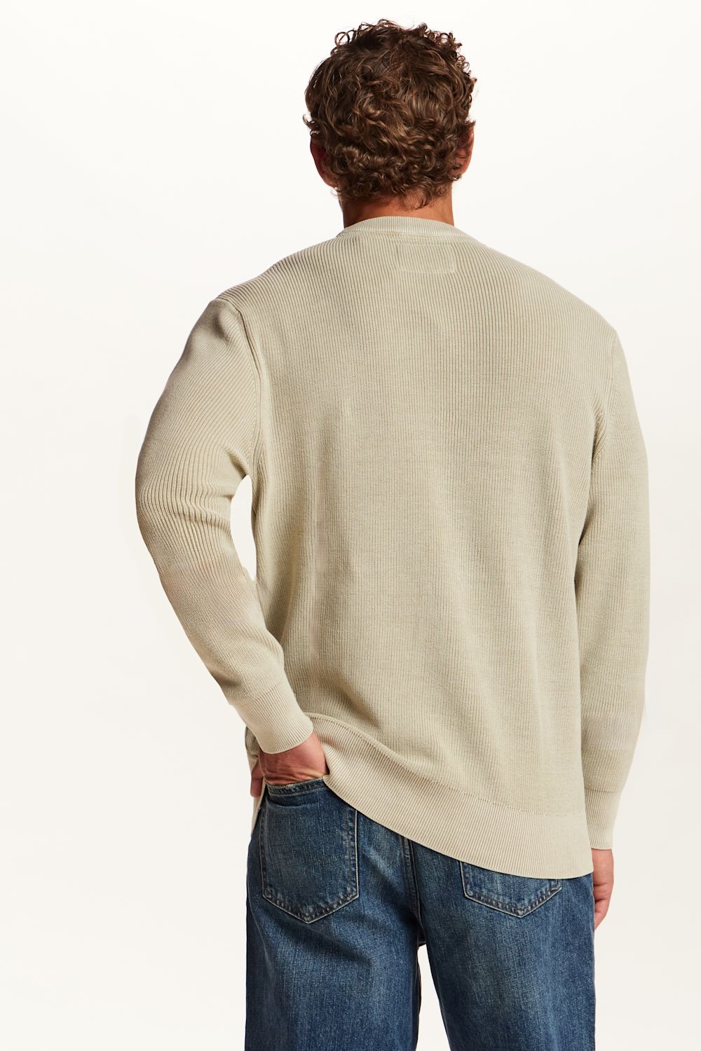 A man viewed from behind wearing a light beige ribbed knit sweater and blue jeans against a plain white background.