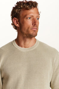 A close-up shot of a man wearing a light beige ribbed knit crewneck sweater against a plain white background.