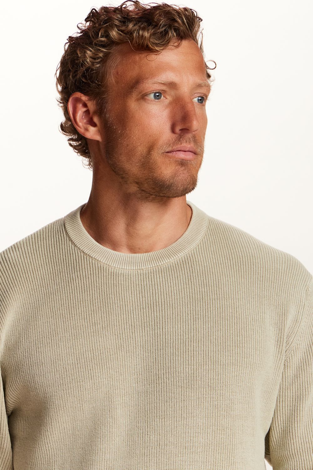 A close-up shot of a man wearing a light beige ribbed knit crewneck sweater against a plain white background.