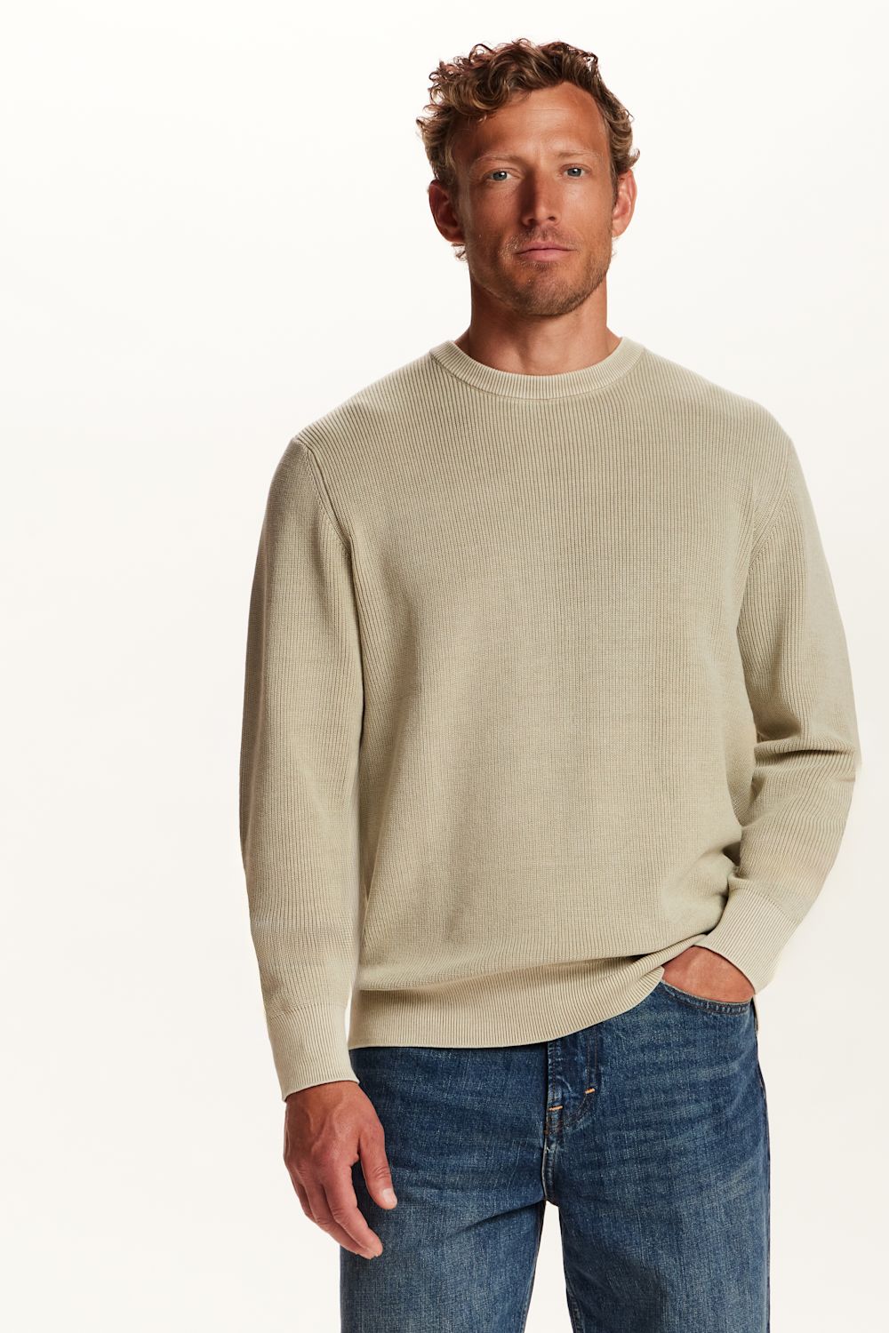 A man wearing a light beige crewneck sweater and blue jeans, standing against a plain white studio background.