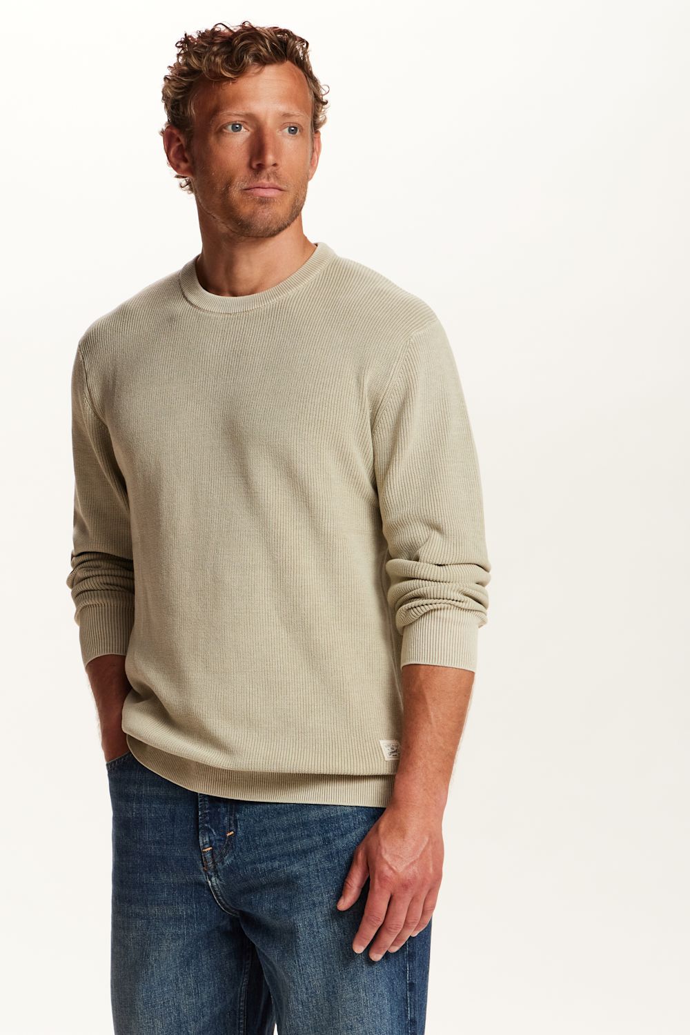 A man wearing a light beige crewneck sweater and blue jeans, standing against a plain white studio background.