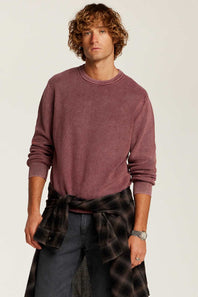 A man wearing a washed, heathered dusty rose crewneck sweatshirt, dark gray jeans, and a dark plaid shirt tied around his waist, photographed against a light off-white studio background.