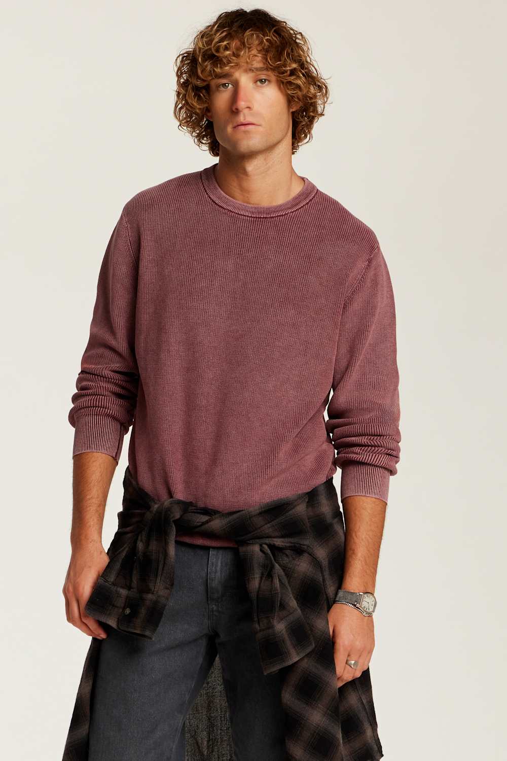 A man wearing a washed, heathered dusty rose crewneck sweatshirt, dark gray jeans, and a dark plaid shirt tied around his waist, photographed against a light off-white studio background.