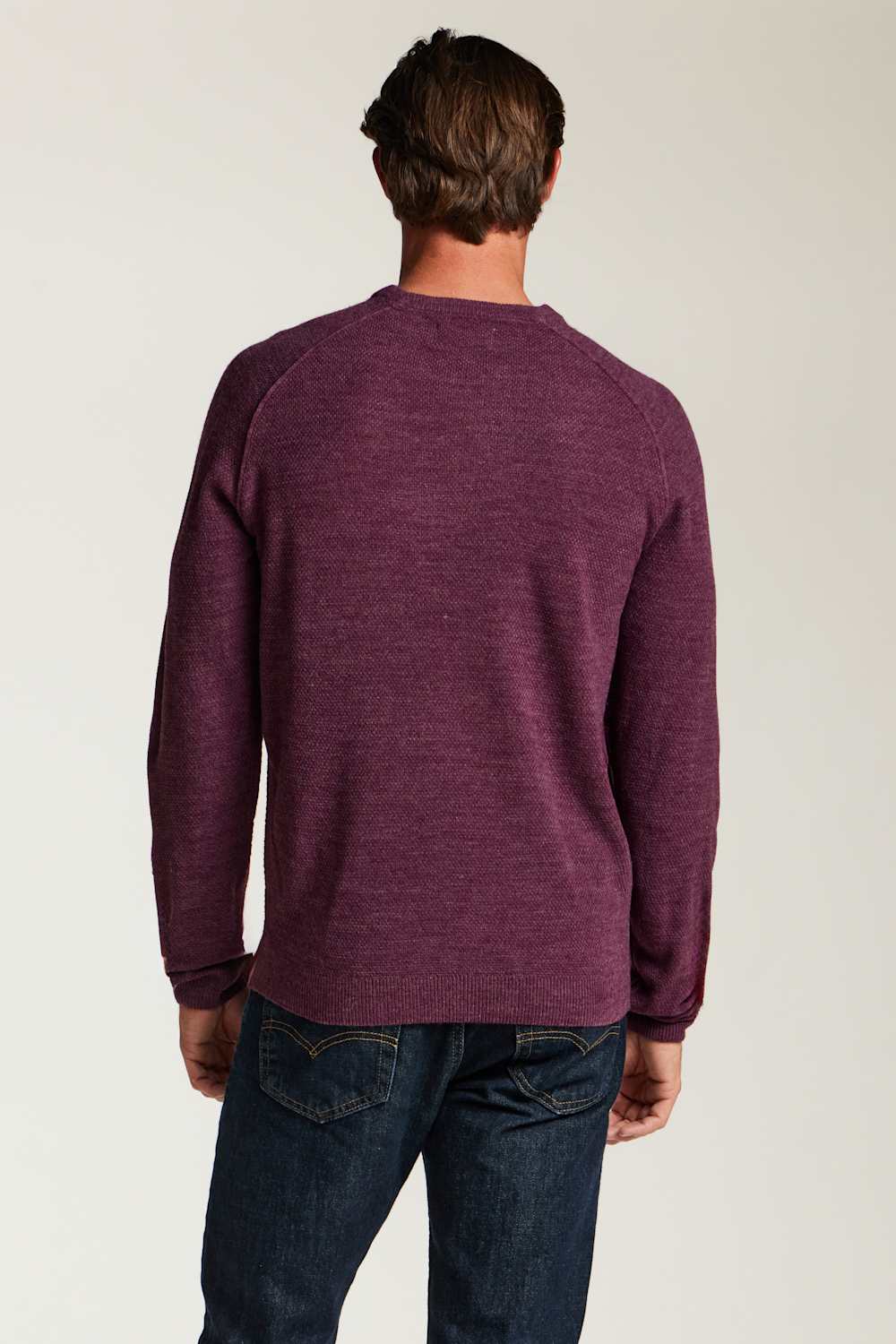 A man viewed from behind wearing a heathered dark purple long-sleeve crewneck sweater and dark blue jeans, standing against a plain light gray studio background.