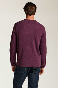 A man viewed from behind wearing a heathered dark purple long-sleeve crewneck sweater and dark blue jeans, standing against a plain light gray studio background.