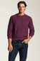 A man wearing a heathered purple long-sleeve crewneck sweater and dark blue jeans, standing against a light beige studio background.