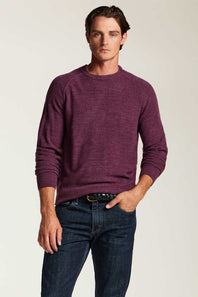 A man wearing a heathered purple long-sleeve crewneck sweater and dark blue jeans, standing against a light beige studio background.