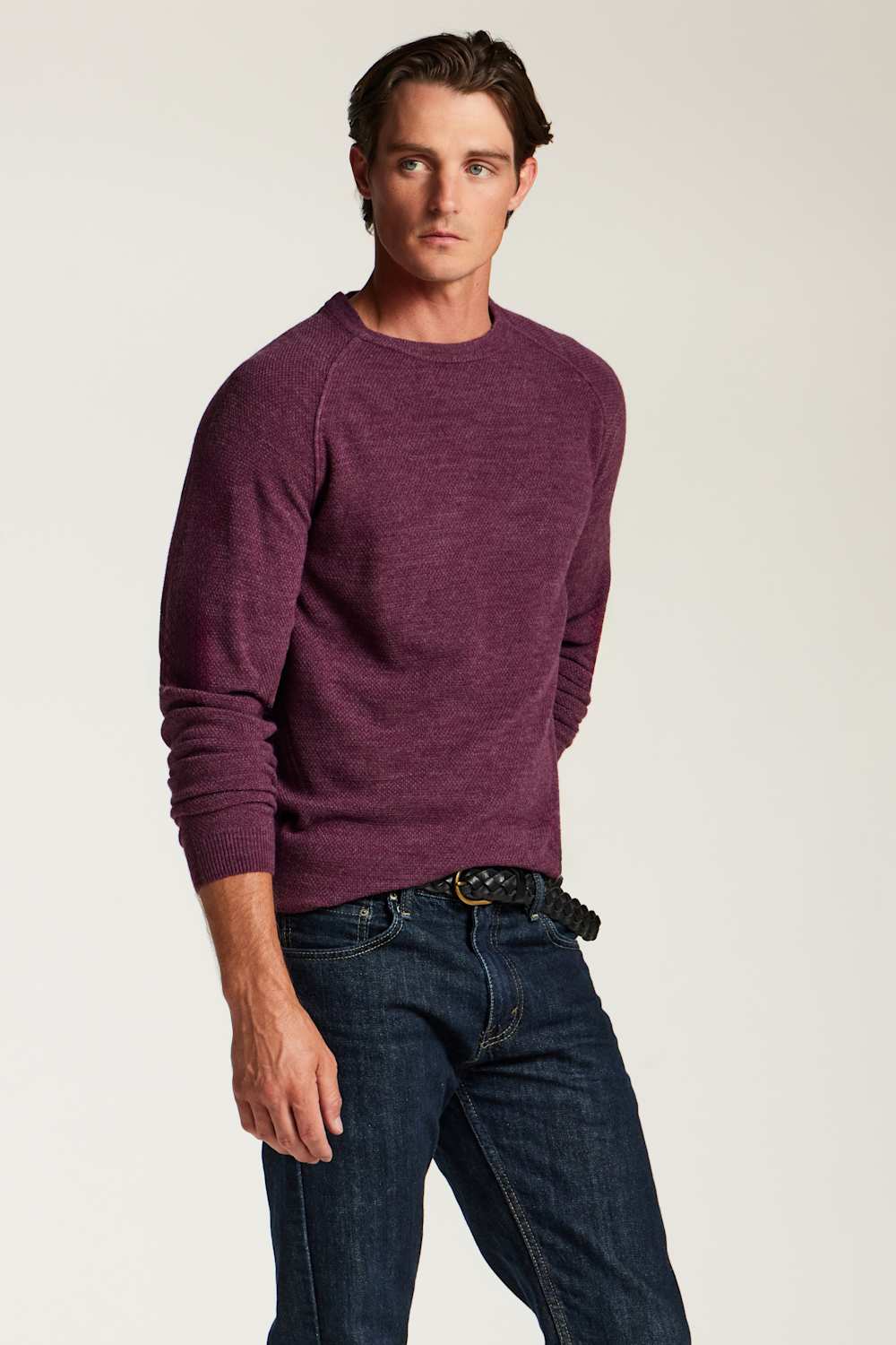 A man wearing a heathered plum-purple long-sleeve knit sweater and dark blue jeans, standing against a plain light gray studio background.