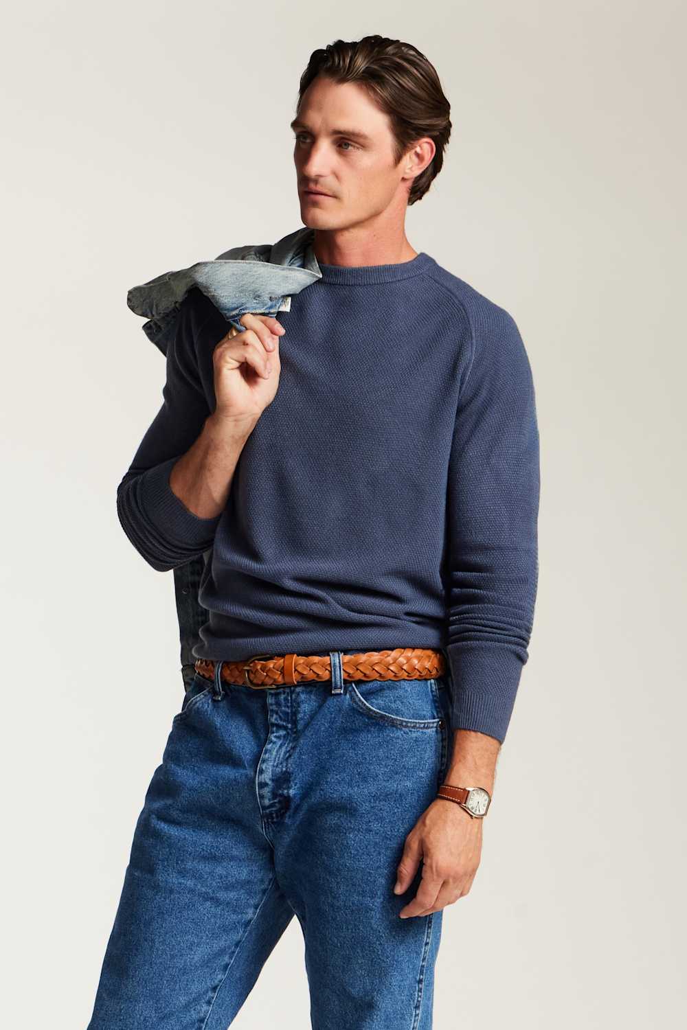 A man wearing a navy blue crewneck sweater, blue jeans, and a brown braided belt, holding a light blue denim jacket over his shoulder against a light gray studio background.
