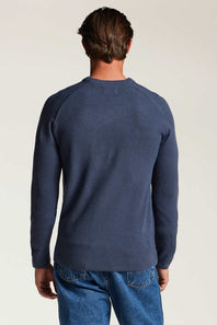 A man viewed from behind wearing a navy blue long-sleeve crewneck sweater and blue jeans against a plain light background.